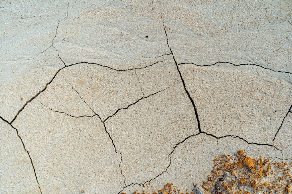 A textured full-frame shot reveals an expanse of dry, cracked earth. The beige and brown earth shows many fissures.