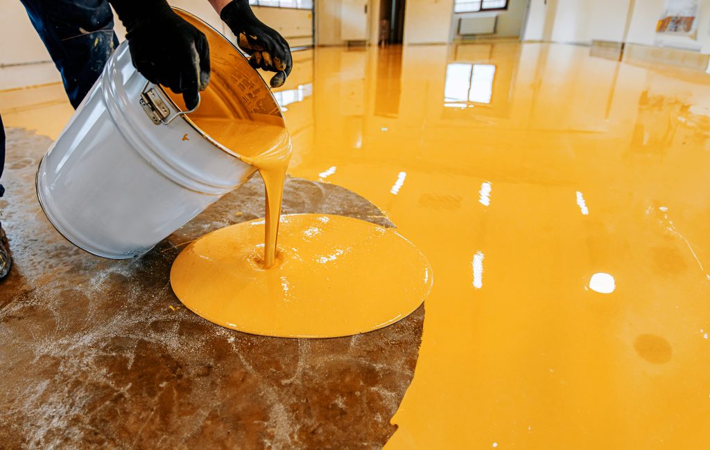 An industrial floor is being coated with orange epoxy in this interior shot, located in South West Virginia. The gloved hands of an adult hold a tilted, light gray bucket, from which a stream of vivid orange liquid flows onto the floor. 